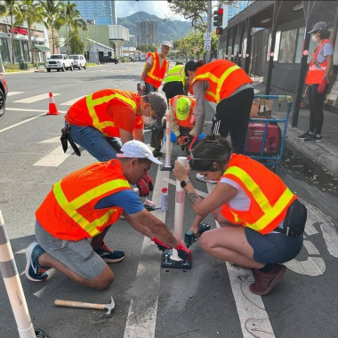 Group of people in high-visibility vests working on a flex post marking the bike lane with tools and equipment. There’s a cone marking the adjacent car lane as closed. 