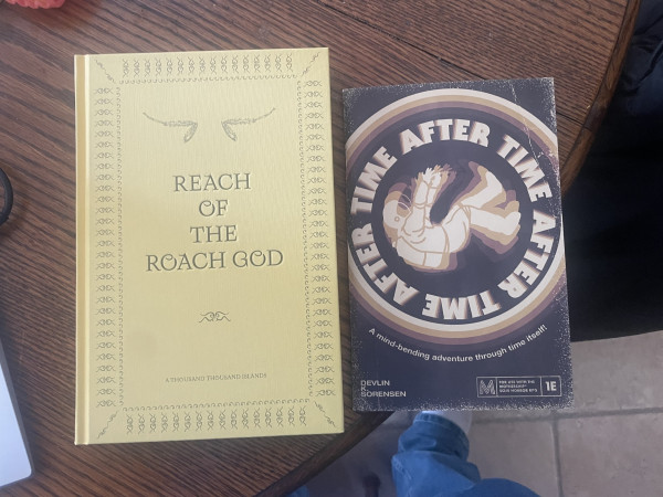 Two books on a wooden surface. The left book is titled "Reach of the Roach God" with a yellow cover, and the right book is titled "Time After Time After Time" with a depiction of an astronaut falling through a circular design.