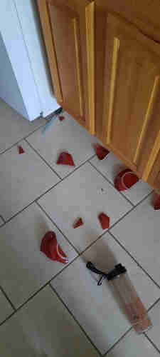 A shattered, red, ceramic bowl and a dropped plastic smoothie bottle on a light colored kitchen floor. Brown cabinets and a white fridge can be seen at the top of the photo