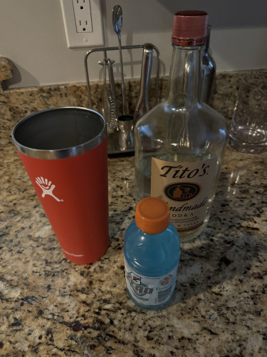 An insulated cup with a small bottle of Gatorade and a large bottle of vodka on a counter. 