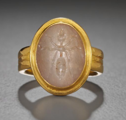 A gold ring with a white stone. An ant, as seen from above is carved into the stone. 