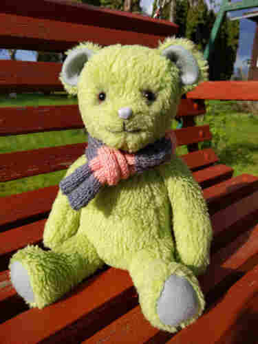 Cute teddy bear sitting on a bench in the garden. The face has shiny eyes, a patterned button nose and a happy smile. The fur is green and fluffy with grey felt ears and paws, wearing a crocheted scarf from pink and grey wool.