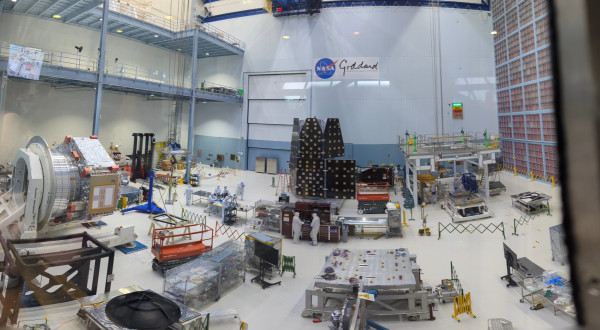 A panorama shows the NASA Goddard clean room, a large, 4-story white room. In the room are several metal tables, testing equipment, scissor lifts, and parts of the telescope. The NASA Goddard logo is on the back wall. Several people are working, dressed head to toe in "bunny suits", white protective suits used to control contamination.