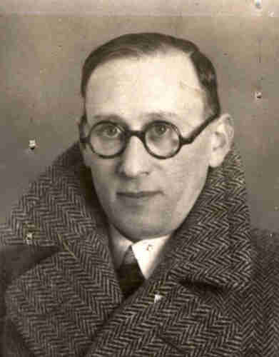 Portrait photograph of a man wearing a coat with very wide lapels against the collar. He is wearing glasses with dark round frames. He has short dark hair combed to the right.