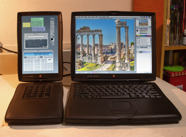 A photo of an apple PowerBook G3 14" laptop from 1998, and its matching PowerPod 3 external screen, keypad, scsi drive and accessory battery. The PowerBook's screen shows Photoshop with some marble columns and other aesthetic type stuff, while the PowerPod shows Photoshop's tool palette, some sticky notes, a keyboard viewer and PCalc 2