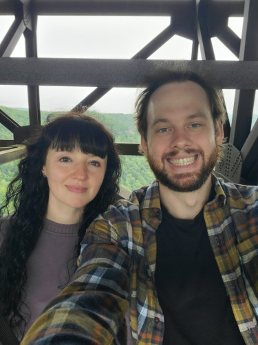 My girlfriend (long dark hair, purple sweater) and me (black tshirt and flannel) standing under a bridge with many trees in the background.