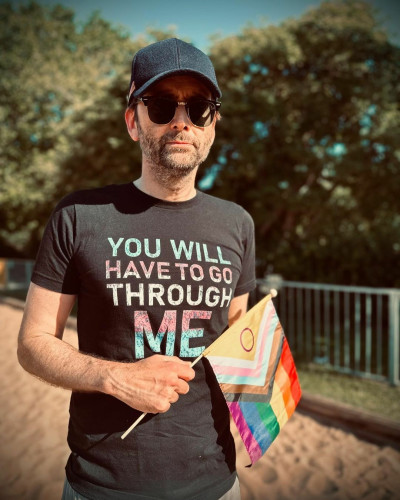 Photo of David Tennant holding the LGBTQ flag, wearing a black shirt that reads: You will have to go through me (the printing is in pale blue, pale pink, and white)