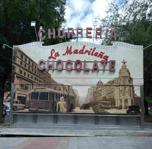Photo of a closed Churrería stall, which is strange as it's normally open.
It is large, the size of a small shop on the side of the road and the shutters has an art mural of urban Madrid with a tram going across the street, a building behind it, and the Metropolis building which is a famous icon in Madrid.
Above in lights that are turned off are the words Churrería, chocolate and the name of the place, La Madrileña
