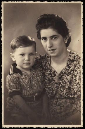 Vintage black and white photograph of an adult and a young child. The adult is sitting and wearing a floral patterned dress, while the child, seated on the adult's lap, is dressed in a striped outfit. Both are looking directly at the camera. The background is neutral.