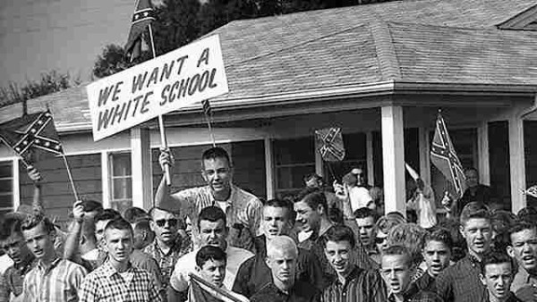 A group of white high school aged boys in a black and white photo from the Civil Rights era. There are several confederate flags and a sign reading: We Want a White Scool