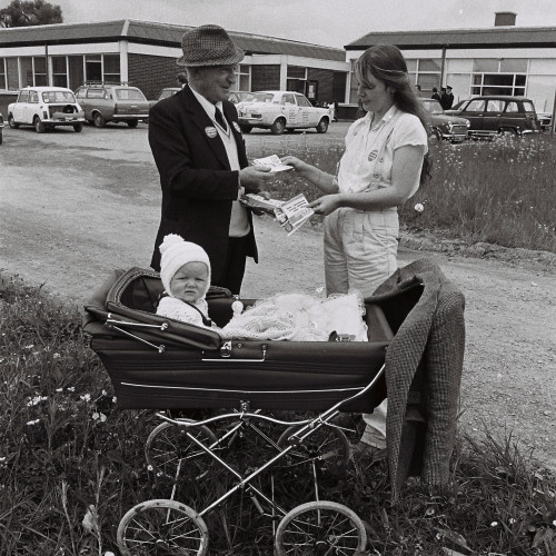 A black and white photo of a campaigner passing an informational leaflet to a mother with her baby in a pram. 