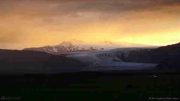 A photo of a mountain range with a curving glacier descending from it. The sun is setting, and the scene is tinted by yellow and orange light. The mountains around it and nearer to ground level are dark and no details can be seen clearly except for a car's headlights visible on the bottom right.