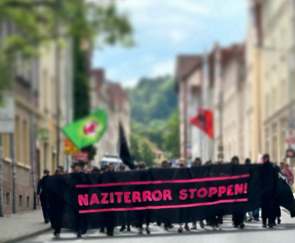 Demonstration. Man sieht verschwommen mehrere Menschen hinter einem schwarzen großen Transparent auf dem in pinken Großbuchstaben steht „Naziterror stoppen!“ im Hintergrund wehen zwei Antifa-Fahnen