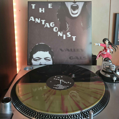 A Gold & Smoke Translucent Split With Red Splatter vinyl record sits on a turntable. Behind the turntable, a vinyl album outer sleeve is displayed. The front cover shows 2 halves of faces. To the top right of the cover is the lower half of a woman's face screaming.  The bottom left of the cover is the top half of another persons face.
