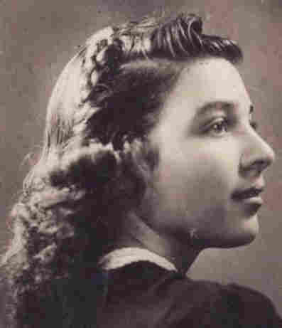 A photo of the face and shoulders of a young woman. She is photographed from the back and she turns her head to show her profile. She has ling hair styled waves and curls. 