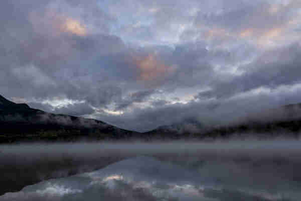 A mountain range at sunrise stands out black against the stormy sky, clouds of grey and grey-lavender torn and gilded with rose, descending so far as to veil the mountains’ flanks. Below, a line of grey mist separates the horizon from the mirror-still lake below, reflecting back the sky and mountains. 