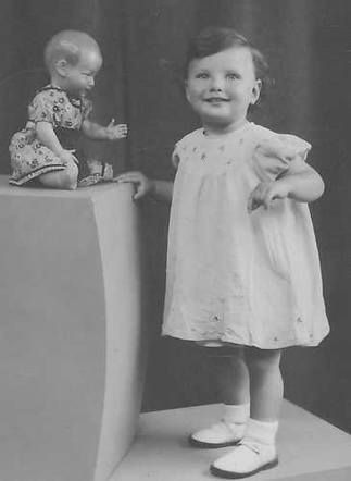 A vintage black-and-white photo of a child standing smiling towards the camera, with a doll seated on a high pedestal beside them in a studio setting.