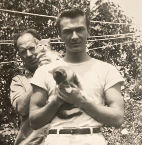 Black and white photo of a muscular young white man with dark, slicked back hair, staring at the camera and holding up a shorthaired tuxedo tabby kitten. An older white man behind him props another kitten up on his shoulder.