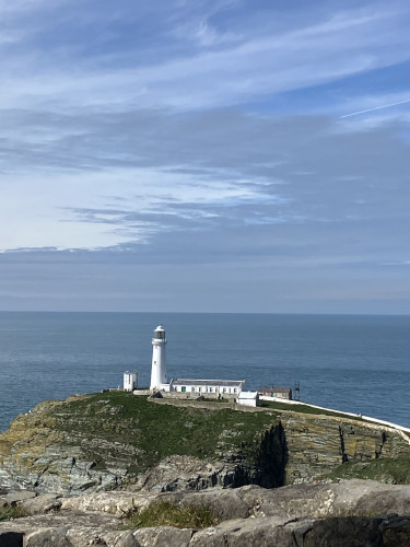 A white lighthouse (South Stack Lighthouse / Ynys Lawd) and attached building situated on a grassy cliff edge, overlooking a calm sea at Anglesey with a partly cloudy sky above.