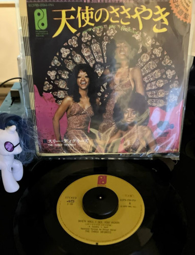 A 7" record sleeve on a stand above a 45rpm record on a turntable. The sleeve as a title at the top 「天使のささやき」Below are three women in gold dresses sitting before a round stained glass window.

Below is a record with a dark yellow label with a stylized magenta P logo. Below the spindle hole is title "When Will I See You Again"