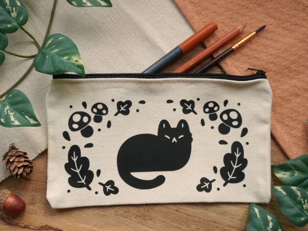 A photograph of a canvas pouch with a black print on it of a cat and autumn decorations 