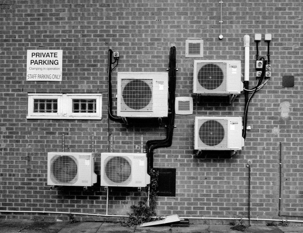 Black and white photo showing part of the back wall of some shops. There are five air conditioning units and several vents set in the brick wall, plus a sign reading "PRIVATE PARKING, Clamping in operation, STAFF PARKING ONLY".