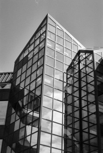 A glass and steel building soars to a sharp point in this portrait format black and white photo. The main part appears slightly off vertical in the frame in this view. There is a lower secondary structure to the right.