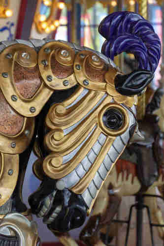 A black carousel horse, richly caparisoned in gold and silver with a purple plume waving from his poll, with glittering clear gems set into his armour along his neck. His head is bent into his chest, teeth bared, and his eye is wide and gleaming. Other carousel horses and lights are visible, blurry, in the background. 