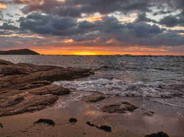 Sunset at a rocky beach with waves crashing onto the shore under a cloud-filled sky. From the setting sun you can only see a bright glow on the horizon, some clouds are colored orange. Boats at anchor sway on the water in the distance.