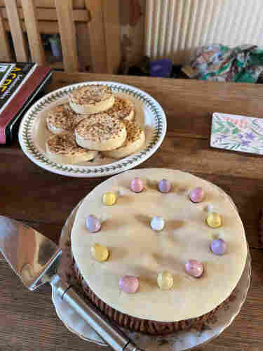 A fruit cake with a layer of icing decorated with mini eggs. Behind the cake is a plate of crumpets!