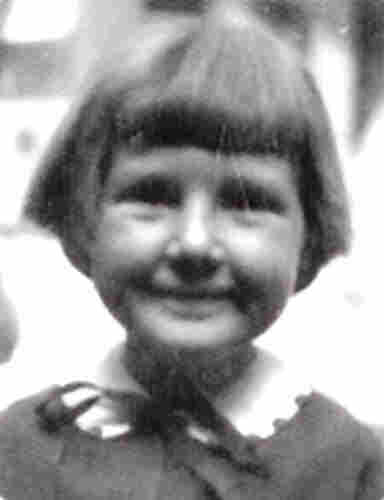 A picture of a smiling girl's face. She has a long fringe evenly cut in the middle of her forehead and her side hair covers her ears. She has a dark ribbon tied under her neck. She is wearing a blouse with a collar. 