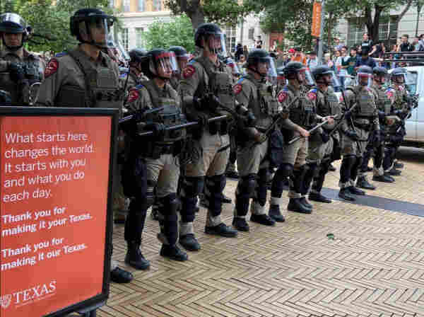 Riot police stand in a line next to a sign that says "What starts here changes the world. It starts with you and what you do each day." 