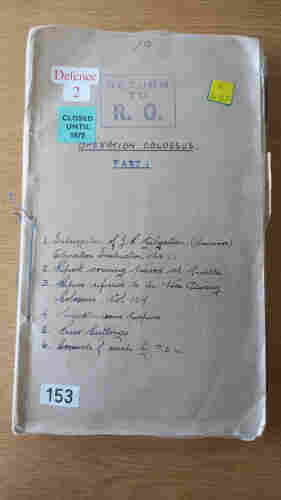 Photo of a historical document with a brown cardboard cover.

Title is "Operation Colossus Part 1". There is a handwritten list underneath:

1. Interrogation of J R Gilgallan (survivor) operation instruction No. 1
2. Report covering period at Malta
3. Papers referred to in War Diary Colossus, vol 107
4. Miscellaneous papers
5. Press cuttings
6. Accounts of escape by P.O.W.

There are several labels: "Defence 2", "Closed until 1972", "E 432", and "153".

"Return to R.O." has been stamped on it.