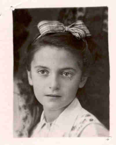 Black and white photograph of a young girl. Her head and shoulders are visible. She is wearing a white shirt with a waistcoat in a floral pattern. The girl has dark hair tied up with a band with a bow. She has a slightly open mouth and a slightly nostalgic expression on her face.