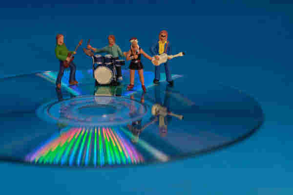Miniature figurines of a band, including a guitarist, drummer, singer, and bassist, positioned on a CD with a reflective rainbow pattern on a blue background.