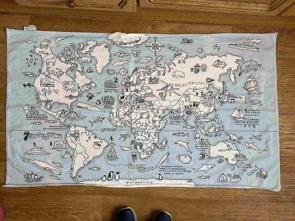 A piece of fabric on the floor. The fabric is covered with a map of the world and some of the countries have animals or landmarks in them - Sydney opera house in Australia, moose and bears in Russia etc. the sea is blue and the countries are other things are white. 