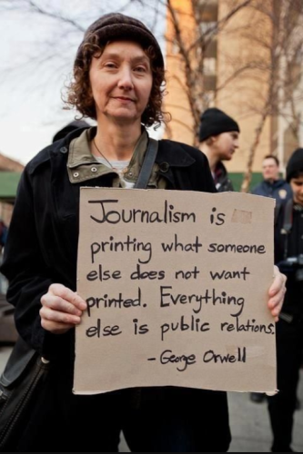 Photo: middle-aged woman attending a protest rally while holding a cardboard sign printed in black ink with a quote attributed to George Orwell: “Journalism is printing what someone else does not want printed: everything else is public relations.”

source: https://www.canyoncountryzephyr.com/blog/2012/06/13/george-orwell-on-journalism/

