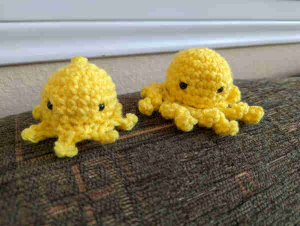 Two crochet yellow octopi with black eyes, one with stumpy legs, the other with longer twisted legs. They both kinda look mad.