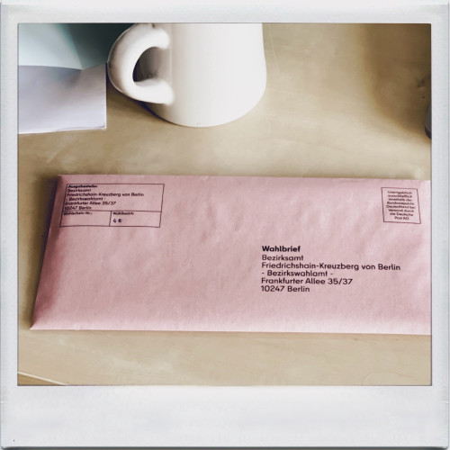 A pink election letter (Wahlbrief) addressed to Bezirksamt Friedrichshain-Kreuzberg von Berlin lies on a table next to a white coffee mug.