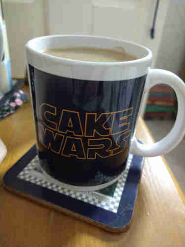 Black mug of tea on a bedside cabinet, with a white handle to the right, and the words 'cake wars' in orange text 