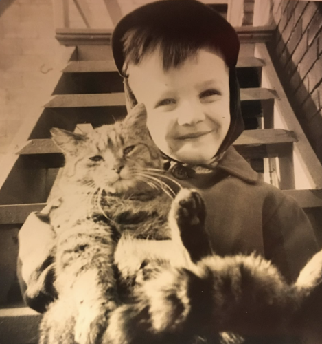 Sepia toned black and white photo of a young white boy smiling at the camera with a look of genuine, radiant joy. In his arms he cradles a shorthaired tabby cat upside down like a baby. The cat looks perfectly comfortable and content.
