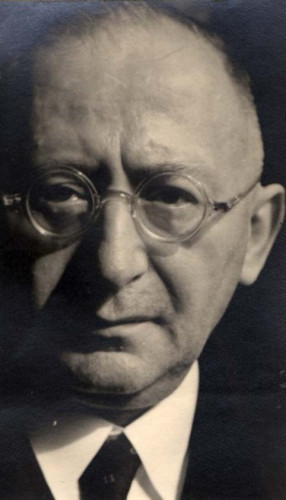 Close-up portrait of a mature man wearing round glasses, displaying a serious expression.