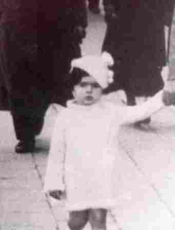 A little girl in a white short dress photographed on the sidewalk. She is holding someone's hand. On her head is a small ornate hat.