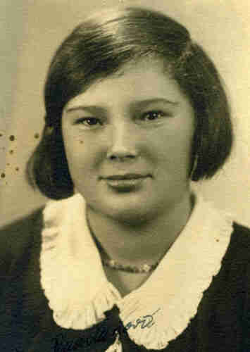 Portrait photograph of a young woman. She has straight hair reaching behind her ears. She has a round face. She is smiling slightly. She is wearing a dark blouse with a white wide collar. There are beads around her neck.