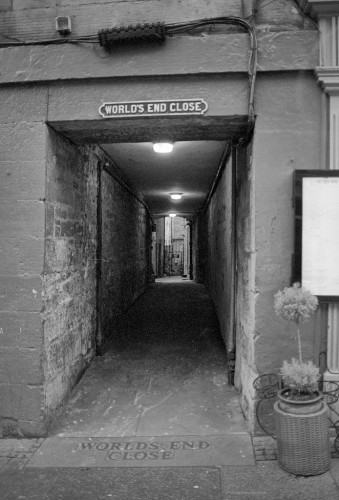 Portrait format black and white photo of a passageway in a stone-built building, with the words "WORLD'S END CLOSE" set above it, and in the stone below. There are 2 lights in the ceiling of the passage, then a gate and some light beyond. To the right a potted plant and a noticeboard are visible, and there is a  massive electrical junction and other wiring above.