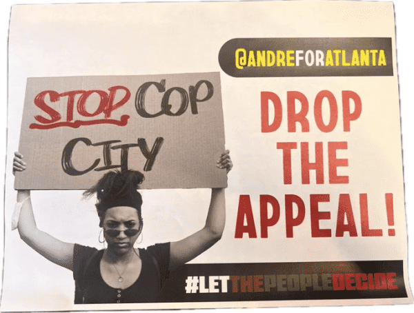 On the left side of the picture there is a protester holding a sign reading "STOP COP CITY" above their head.  There is text on the right side of the picture reading "@andreforatlanta DROP THE APPEAL! #LetThePeopleDecide"