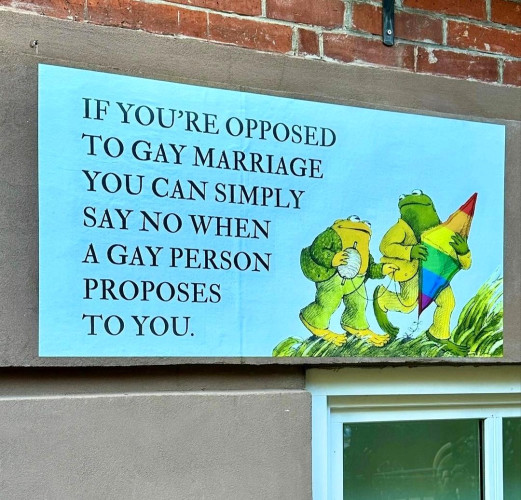 Streetartwall. A blue poster with dark blue text was stuck to a brown house wall with a window. Two drawn green frogs carry a rainbow-colored stunt kite over a green meadow.
"Next to it is the following text: 
If you're opposed to Gay Marriage you can simply say no when a gay person proposes to you."
Info: WestvillageFamous is an anonymous designer who sticks signs with mostly political and satirical messages on the wall of his house in New York.