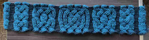 A piece of crochet fabric for the front of a sweater. The pattern has cables in turquoise against a dark blue/black background.