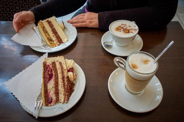 We are looking at a table in a café. In front of us is a plate of Black Forest gateau and a latte macchiato. A person is sitting across from us. In front of her is a plate of nut gateau and a café au lait.