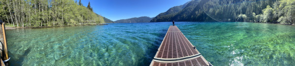 A panoramic shot looking down the pier next to the boat launch on Lake Crescent. The water is vivid green on both sides and blue straight ahead, with light ripples from the bright sun,  and there are mountains all around, and blue sky above. 